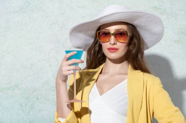 Moda mankeni, beyaz şapka ve güneş gözlüğünü sarı ceketle giyiyor, güneşli bir yaz gününde bir duvarın yanında dinleniyor ve pembe galslerde cyan kokteyli içiyor.
