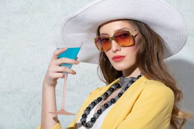 Sarı ceketli, şapkalı, güneşli bir yaz gününde rahatça kokteyl içen genç bir manken. Güneş gözlükleriyle kameraya bakıyor.