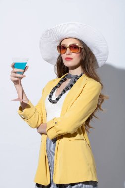 Şık bir manken, sarı ceketli ve şapkalı, güneşli bir yaz gününde beyaza poz veren, güneş gözlüklü bir kokteyl içiyor.