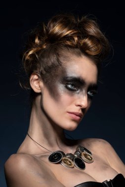 Çekici, yaratıcı makyaj, çok moda portresi, koyu mavi arka plan ve haistyle, geri dönüşümlü malzemeden yapılmış kolye takıyor.