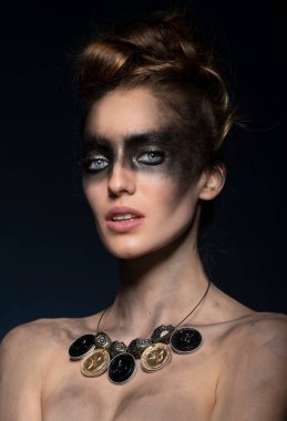 Geri dönüşümlü malzemeden yapılmış kolye takan ve yaratıcı kirli makyajlı genç bir kızın harika bir portresi. Mavi gözleriyle kameraya bakıyor.