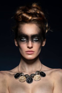 Moda mankeni genç kız, koyu mavi arka planda, kameraya bakıyor, yaratıcı bir makyajla, geri dönüşümlü malzemeden yapılmış saç stili ve kolyesi var.