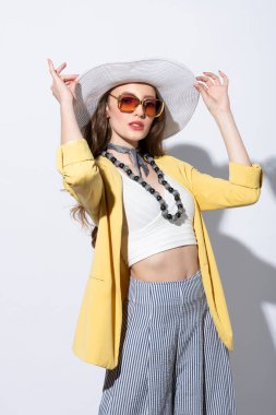 Güzel bir kadın yaz partisine hazır, sarı ceketli, beyaz şapkalı, güneş gözlüğü takıyor, güneş ışığının altında beyaz üzerinde poz veriyor.