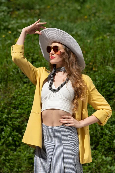 魅力的な女性は エレガントな夏のドレスで 緑の穀物の近くで 彼女は白い帽子で 黄色のジャケットとサングラスを着ていて 日光浴をしています — ストック写真