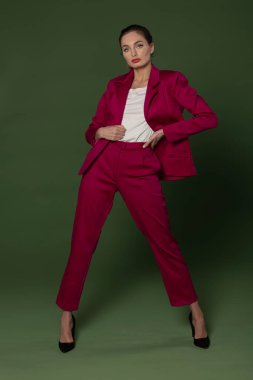 Fuşya kıyafeti, ceket ve pantolonla poz veren çarpıcı bir manken, yeşilde poz veriyor.