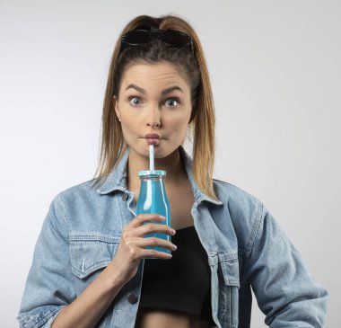 Komik, genç, beyaz bir kız, elinde bir şişe mavi içecekle, sıradan bir ceket giymiş, kameraya bakıp surat yapıyor.