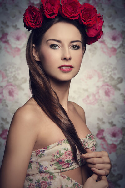 Close-up portrait of lovely girl with long smooth brown hair and sprig dress posing with red roses on the head and natural make-up