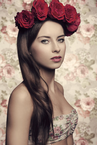 Beauty brunette female posing in spring close-up portrait with long smooth hair, floral dress and cute red roses on her head. Natural make-up, looking in camera