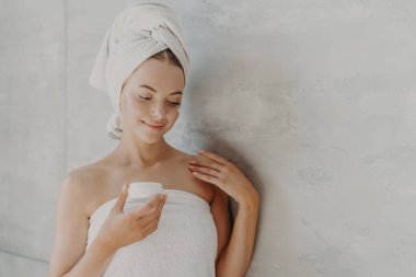Sağlıklı ciltli, makyajı az, yüz kremi sürmüş, duş aldıktan sonra gri arka planda banyo havlusuna sarılmış güzel bir kadın. Sauna sabahı rutini, cilt bakımı konsepti