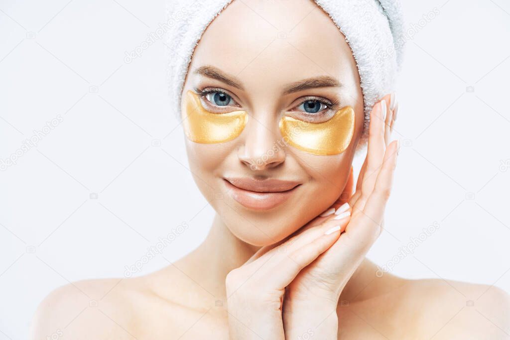 Close up portrait of attractive woman has healthy skin, blue eyes, natural makeup, touches jawline, looks with gentle calm expression, wears bath towel on head, stands shirtless against white wall