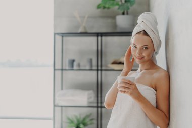 Memnun çekici kadın doğal güzelliğe sahip, pürüzsüz ve sağlıklı bir teni var, yüzüne dokunuyor, spa ameliyatlarından hoşlanıyor, kafasına ve vücuduna beyaz banyo havlusu takıyor, çay içiyor, rahat bir banyoda poz veriyor.