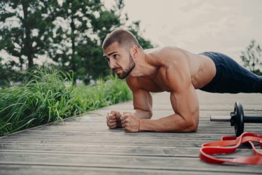 Kas geliştirici tahta duruşunda duruyor. Dayanıklılık gösteriyor ki güçlü pazılara sahip. Açık havada spor yapıyor, dışarıda poz veriyor, fiziksel olarak iyi durumda. Şınav çeken, konsantre olmuş bir adam.