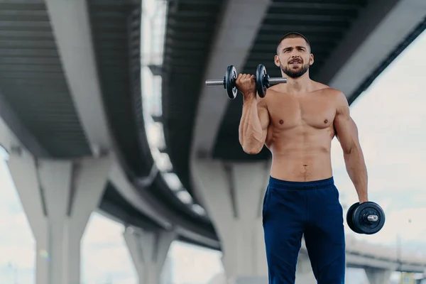 Self confident bearded man exercises with barbells outdoor has naked torso muscular shirtless body trains near bridge does physical heavy weight exercises pumps up arms muscles. Bodybuilding concept