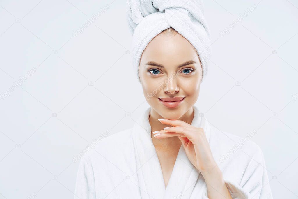 Cropped shot of young good looking European woman looks with calm face, enjoys bath procedures, wears soft white robe, wrapped towel on head, isolated on white background. Morning time, hygiene