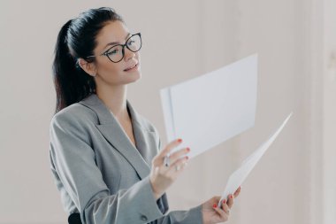 Kadın finans analistinin fotoğrafı yatırım tavsiyeleriyle raporlar hazırlıyor, strateji bilgilerini okuyor, kağıt belgelerle poz veriyor, resmi kıyafetler giyiyor, gözlük takıyor, kapalı yerlerde duruyor.