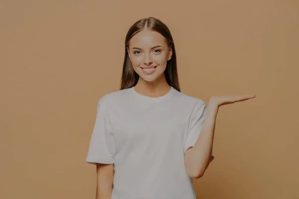 明るい若い女性は 目に見えない物体が茶色の背景に対してカジュアルな白いTシャツのポーズを身に着けていることを示しています 広告宣伝コンセプト — ストック写真