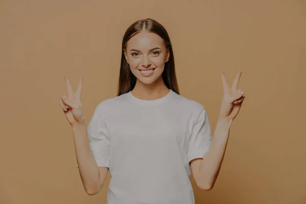 幸せな楽観的な若い白人女性の水平ショット腕を上げる指で平和ジェスチャーを示しています勝利のサインは 数2は茶色の背景に隔離されたカジュアルな白いTシャツを身に着けていると言う — ストック写真