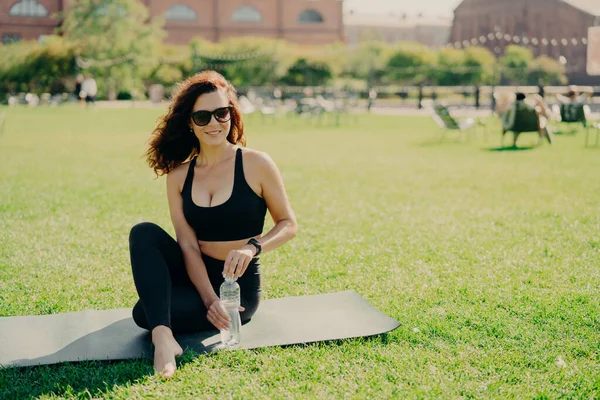 Horizontal shot of active slim woman in sportswear sits on karemat holds bottle of cold water has rest after exercising abdominal muscles poses on green lawn wears sunglasses. Workout at street