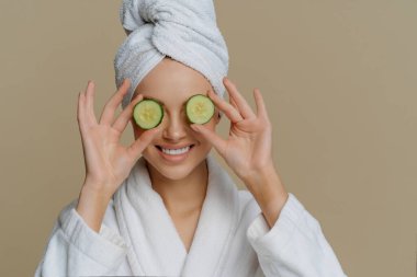 Memnun bir kadının kapalı çekimleri gözlerini salatalık dilimleriyle besler derileri güler, banyo elbisesi giyip havluyla sarmalanır ve iç mekânda durur. Güzellik kaplıcası kozmetoloji konsepti.