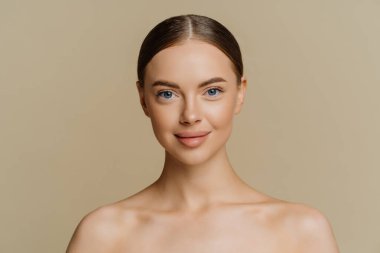 Indoor shot of beautiful young woman with combed hair stands half naked indoor has natural beauty isolated over beige background. Refreshed female model has healthy skin after spa treatments