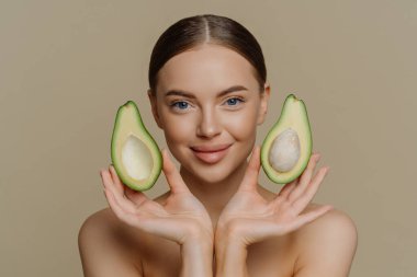 Kişisel bakım ve kaplıca konsepti. Esmer, Avrupalı bir kadının yüzünün yanındaki avokado parçalarını tutuyor. Yüz maskesi öneriyor. Çıplak omuzlar içeride, bej renkli stüdyo arka planında izole edilmiş..