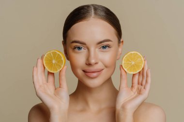 Güzel esmer kadının portresi sulu limon dilimleri tutuyor sağlıklı parlak ciltleri var turunçgillerden vitaminler alıyor kapalı alanda bej arka planda çıplak duruyor. Yüz tedavisi kavramı