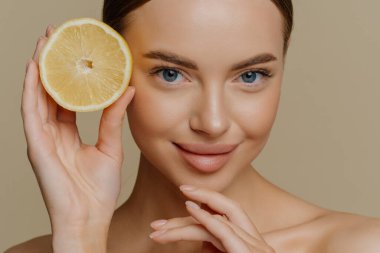 Sağlıklı genç üstsüz esmer kadının güzel portresi çenesine dokunuyor sulu taze limonun yarısını nazikçe tutuyor kahverengi arka planda üstsüz pozlar veriyor. Yüz bakımı kavramı