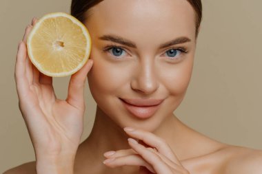 Sağlıklı genç üstsüz esmer kadının güzel portresi çenesine dokunuyor sulu taze limonun yarısını nazikçe tutuyor kahverengi arka planda üstsüz pozlar veriyor. Yüz bakımı kavramı