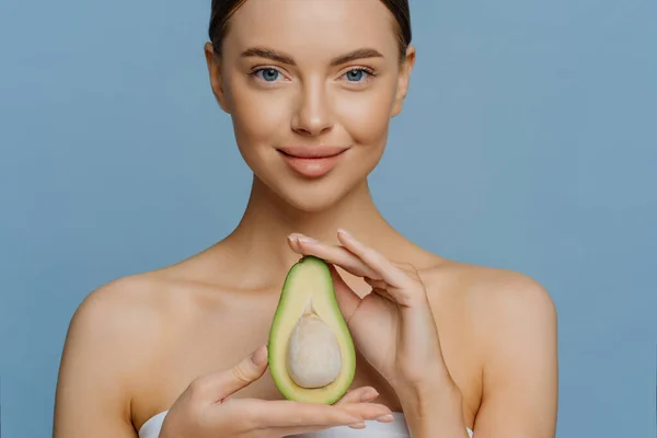 美しい若い白人女性のクロップショットは青いスタジオの背景に隔離された健康的な果物とアボカドのポーズ裸の肩の屋内ポーズの半分を保持しています。皮膚栄養面ケアの概念 — ストック写真