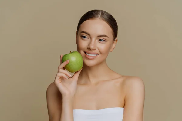魅力的なヨーロッパの女性は顔の近くにリンゴを保持しています笑顔優しく白い完璧な歯を持っています健康的なきれいな肌シャワータオルスタンドに包まれ茶色の背景に裸の肩で. — ストック写真