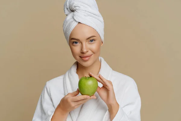 美しい若い女性は頭の上にラップタオルをドレッシングガウンに身を包んだバスを取った後、肌をリフレッシュしている茶色の背景に隔離された多くのビタミンが含まれて緑のリンゴを保持しています。衛生概念 — ストック写真