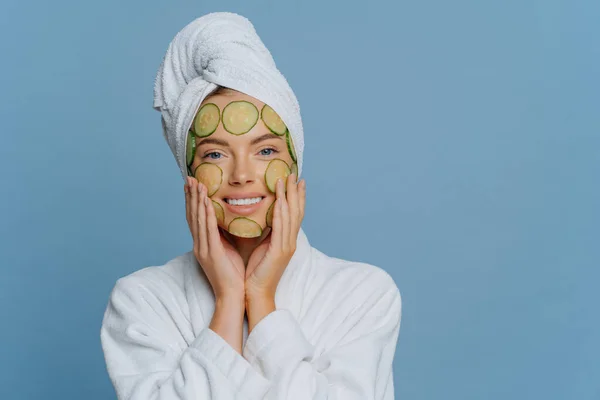 Jovem mulher satisfeita aplica fatias de pepino no rosto sofre procedimentos de beleza faz máscara facial para rejuvenescer a pele vestida com casaco de banho branco e toalha embrulhada na cabeça isolada na parede azul — Fotografia de Stock