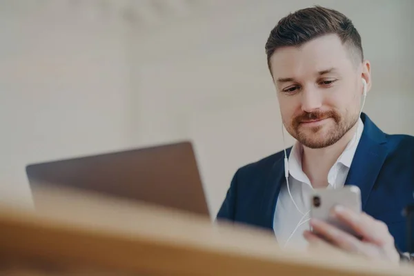 Trabajador de oficina masculino feliz usando auriculares mirando la pantalla del teléfono móvil y viendo video — Foto de Stock