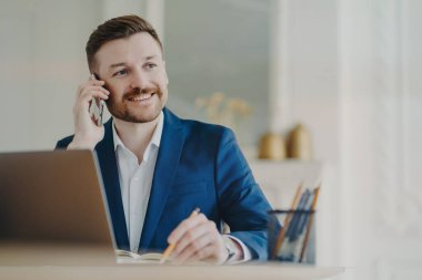 Gülümseyen yakışıklı yönetici dizüstü bilgisayarın önünde telefonla konuşuyor.