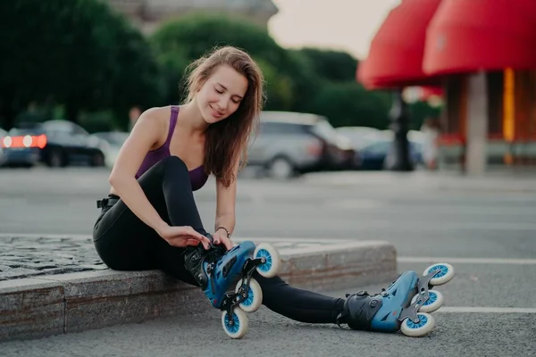 Ευχαριστημένη νεαρή γυναίκα βάζει σε rollerskates πρόκειται να βόλτα rolles σε αστικό μέρος έχει τακτική άσκηση πηγαίνει για επικίνδυνο άθλημα προσαρμόζει κορδόνια — Φωτογραφία Αρχείου