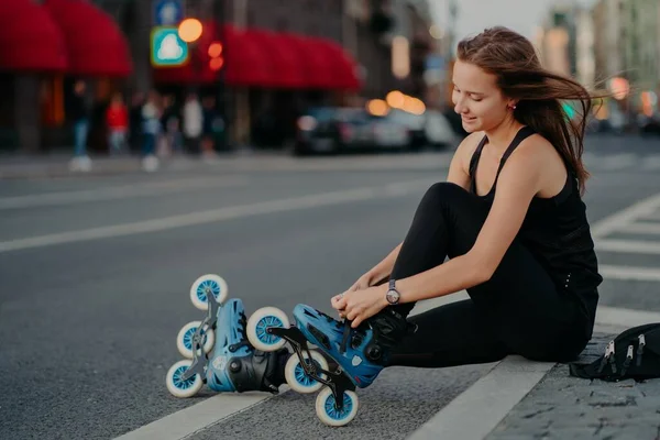 Seitwärtsschuss von aktiver Frau sitzt auf Straße stellt Rollerblades ein bereitet sich auf Skaten vor setzt auf Inlineskates hat bei Wind schwimmende Haare genießt am Wochenende liebstes Hobby — Stockfoto