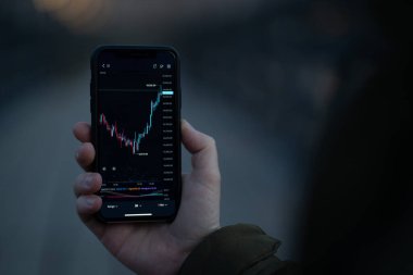 Ekranda gerçek zamanlı Forex grafiği olan akıllı telefonu tutan bir erkek.