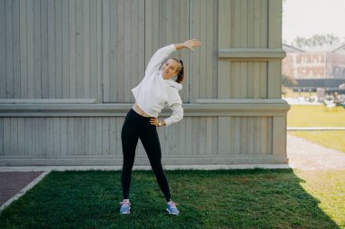 Tam boy aktif sporcu kadın egzersizleri yapıyor. Açık havada vücut egzersizleri yapıyor. Farklı taraflara eğiliyor. Eşofman altı giyiyor ve spor ayakkabıları dinliyor.