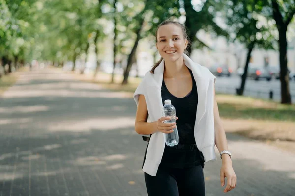 Ao ar livre tiro de mulher desportiva feliz bebe água doce de garrafa tem andar durante o verão verde parque urbano tem sorriso de dente no rosto leva estilo de vida saudável. Restaurar o equilíbrio do aqua após o treinamento — Fotografia de Stock