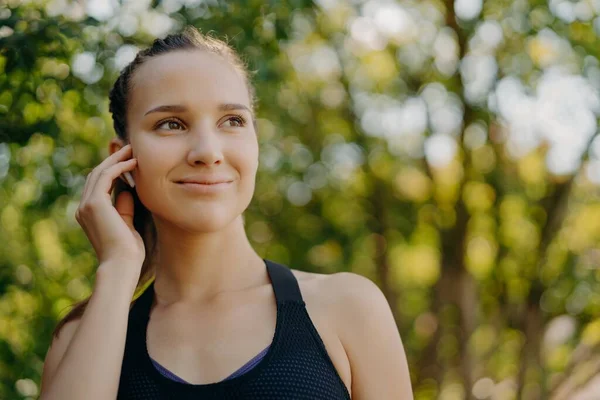 Concept de mode de vie sain. Plan extérieur de la sportive bénéficie playlist préférée dans les écouteurs tandis que l'entraînement dans le parc a sourire doux concentré dans la distance pose sur fond de nature floue — Photo