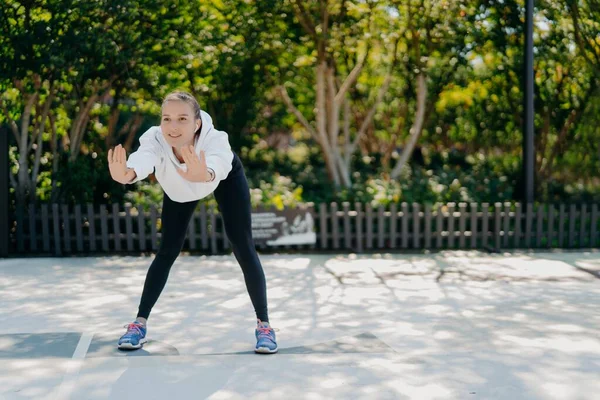 Tiro ao ar livre de mulher desportiva saudável satisfeito inclina-se para a frente mantém as palmas das mãos levantadas faz exercícios em trens ao ar livre no parque se sente bem usa leggings camisola e tênis leva estilo de vida ativo — Fotografia de Stock