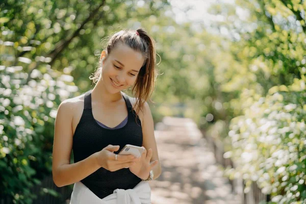 Venkovní záběr šťastný atletický mladá žena používá smartphone sportovní tracker aplikace používá moderní zařízení pro zdraví a fitness je chytrý o tréninku pózuje na rozmazaném přírodním pozadí — Stock fotografie