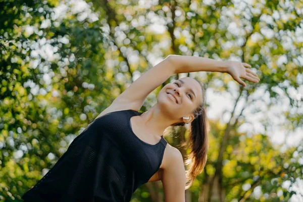 Mulher aptidão desportiva faz aquecimento e alongamento exercícios inclina-se de lado tem expressão rosto conteúdo vestido com poses de desgaste ativo contra fundo da natureza durante o dia de verão. Desporto e motivação — Fotografia de Stock