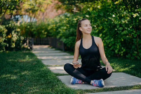 Dark haired young European woman sits crosses legs looks aside with satisfied expression uses smartphone has break after cardio training dressed in active wear and sneaker reads text message — Stock Photo, Image