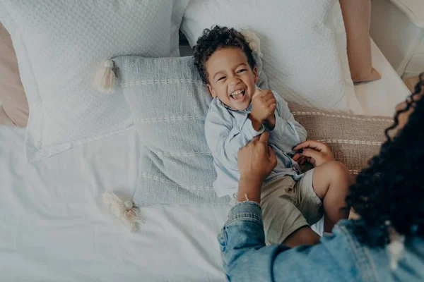 Dulce emocionado mestizo de raza chico acostado en la cama y riendo mientras juega con su amorosa mamá — Foto de Stock