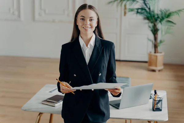 若い笑顔のビジネスマンの女性は、カメラを見て、オフィスの机の前で文書を保持 — ストック写真