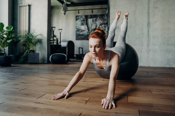 Atletica signora snella con i capelli rossi che esegue esercizi di streching su grande fitball argento durante l'allenamento pilates — Foto Stock