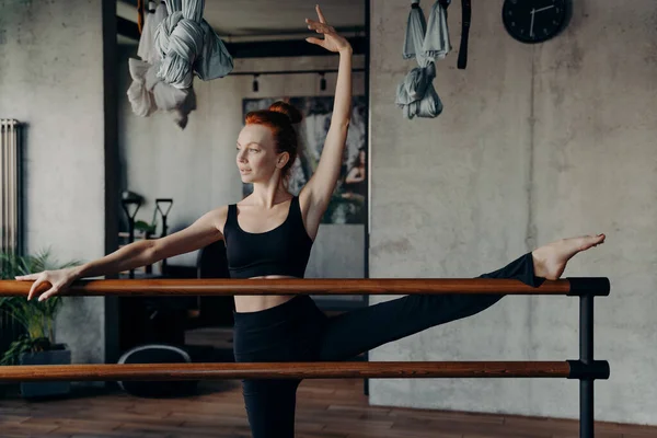 Giovane ballerina dai capelli rossi atletici che allunga la gamba sulla sbarra del balletto in palestra — Foto Stock