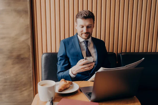 Joven empresario confiado en ropa formal disfrutando del café y trabajando remotamente en la cafetería — Foto de Stock