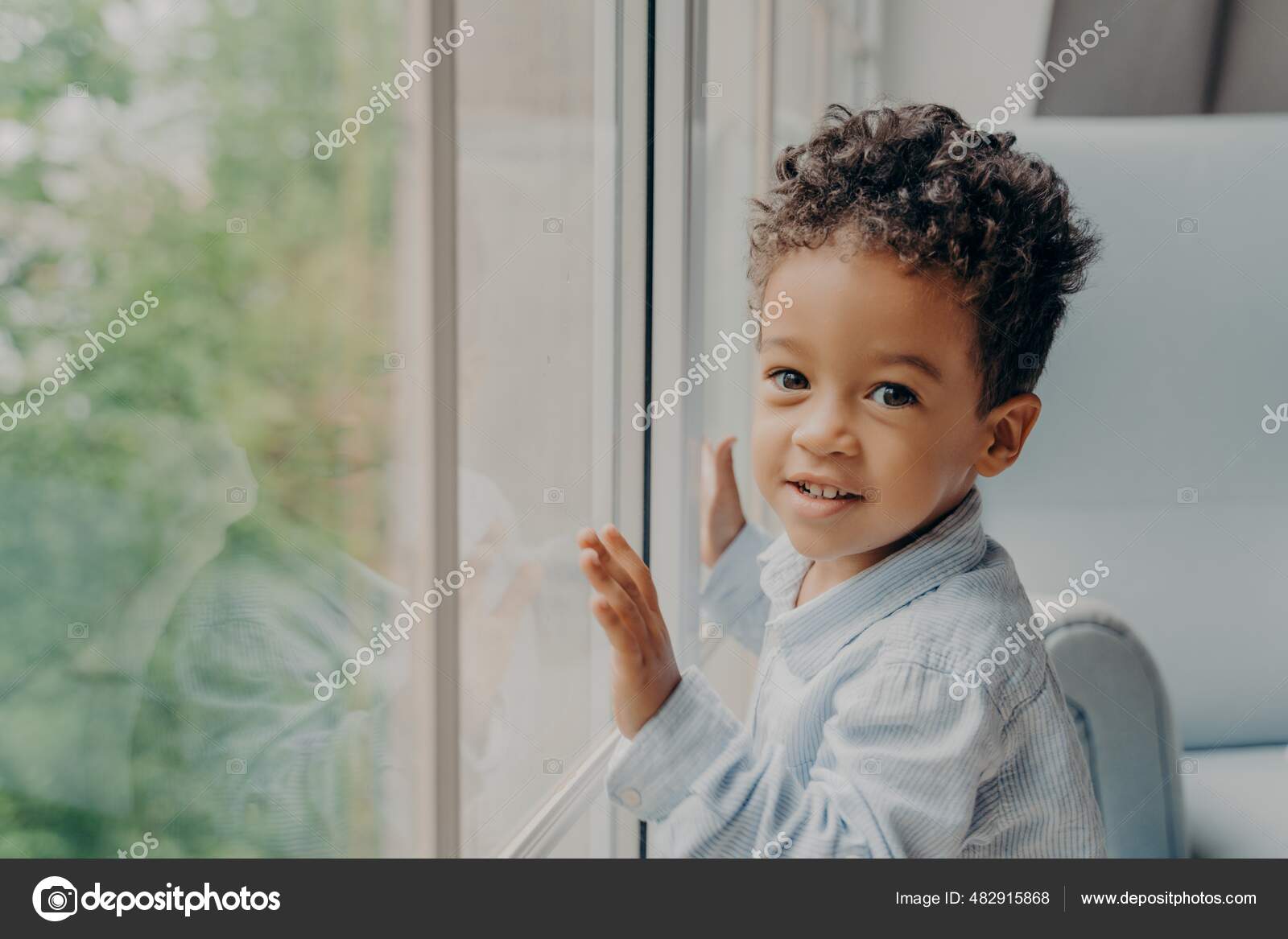 Cute mixed race little curly haired boy in light blue colored shirt next to  large window — Stock Photo © vk_studio #482915868, image size:1600x1167